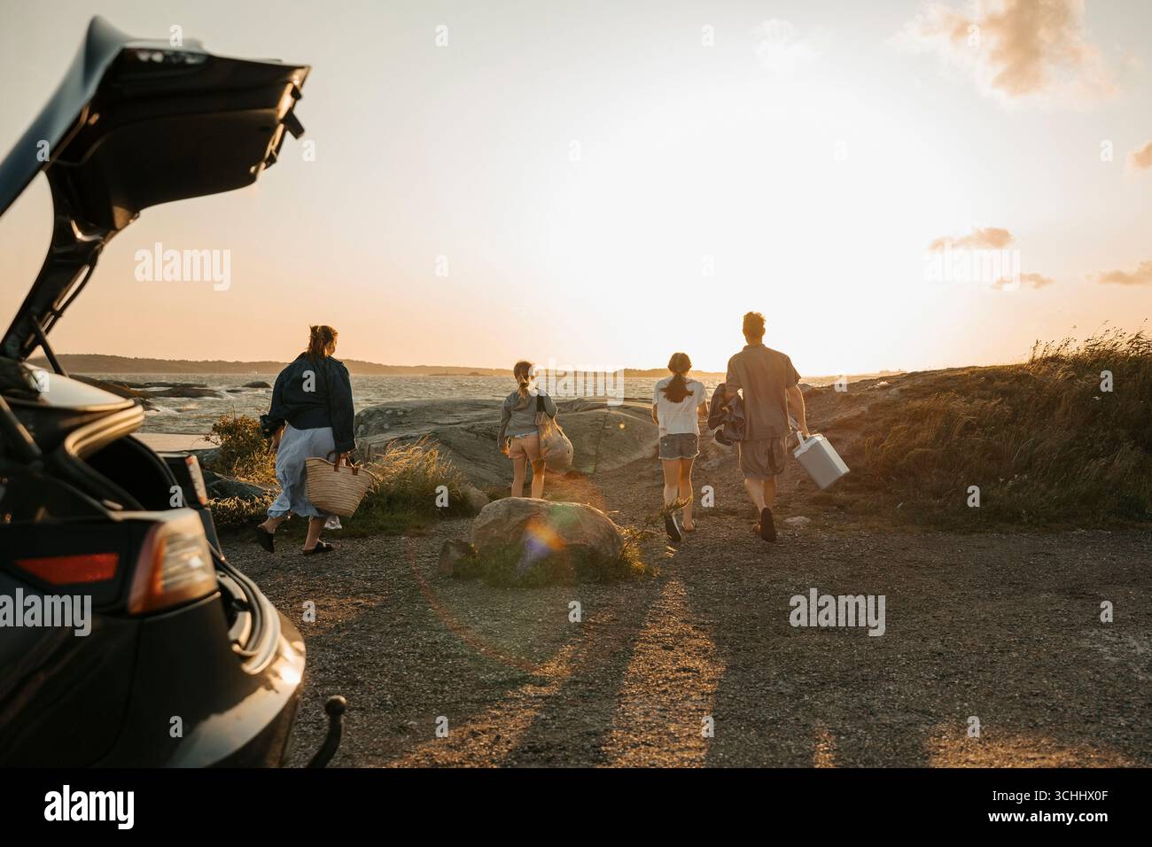 Vista posteriore dei membri della famiglia che camminano verso grandi rocce sulla riva del mare per un picnic Foto Stock