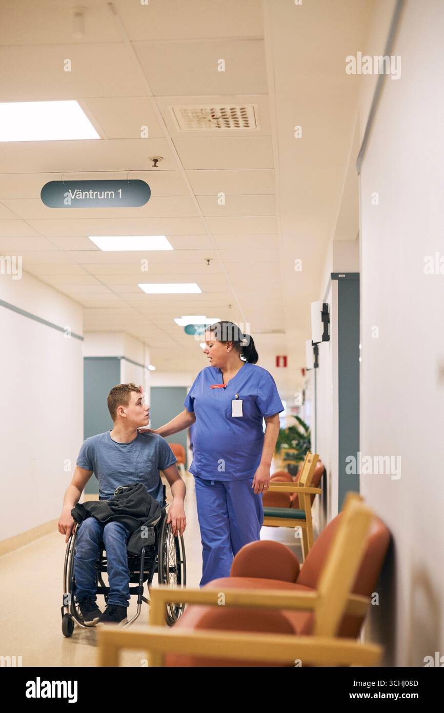 Donna esperta di assistenza sanitaria che cammina con il giovane uomo che gira su sedia a rotelle nel corridoio dell'ospedale Foto Stock