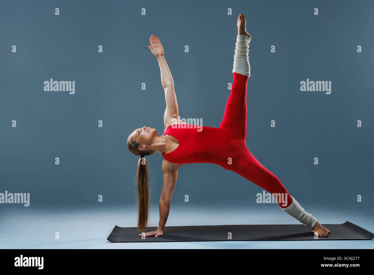Donna atletica in abito rosso in equilibrio con una posa avanzata da yoga con tavolino laterale, mostrando forza e controllo del nucleo. Foto Stock