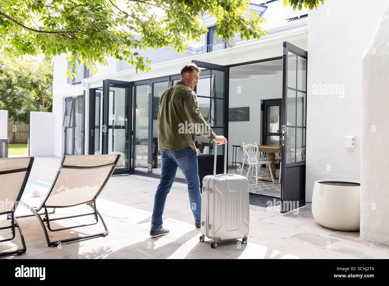 Uomo con valigia che entra in casa moderna attraverso porte di vetro, pronto per il viaggio Foto Stock