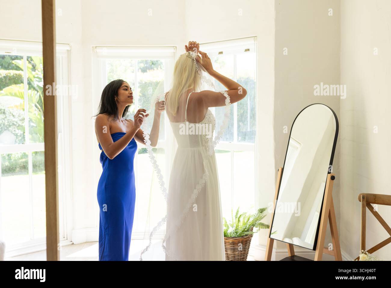 Velo regolabile per la sposa con la damigella d'onore in abito blu che aiuta vicino a un grande specchio Foto Stock
