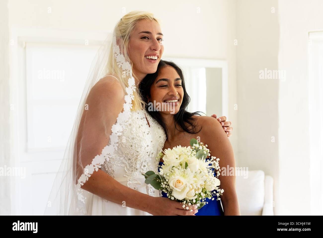 Abbracciando la damigella d'onore, la sposa regge il bouquet, entrambi sorridenti in una camera luminosa Foto Stock
