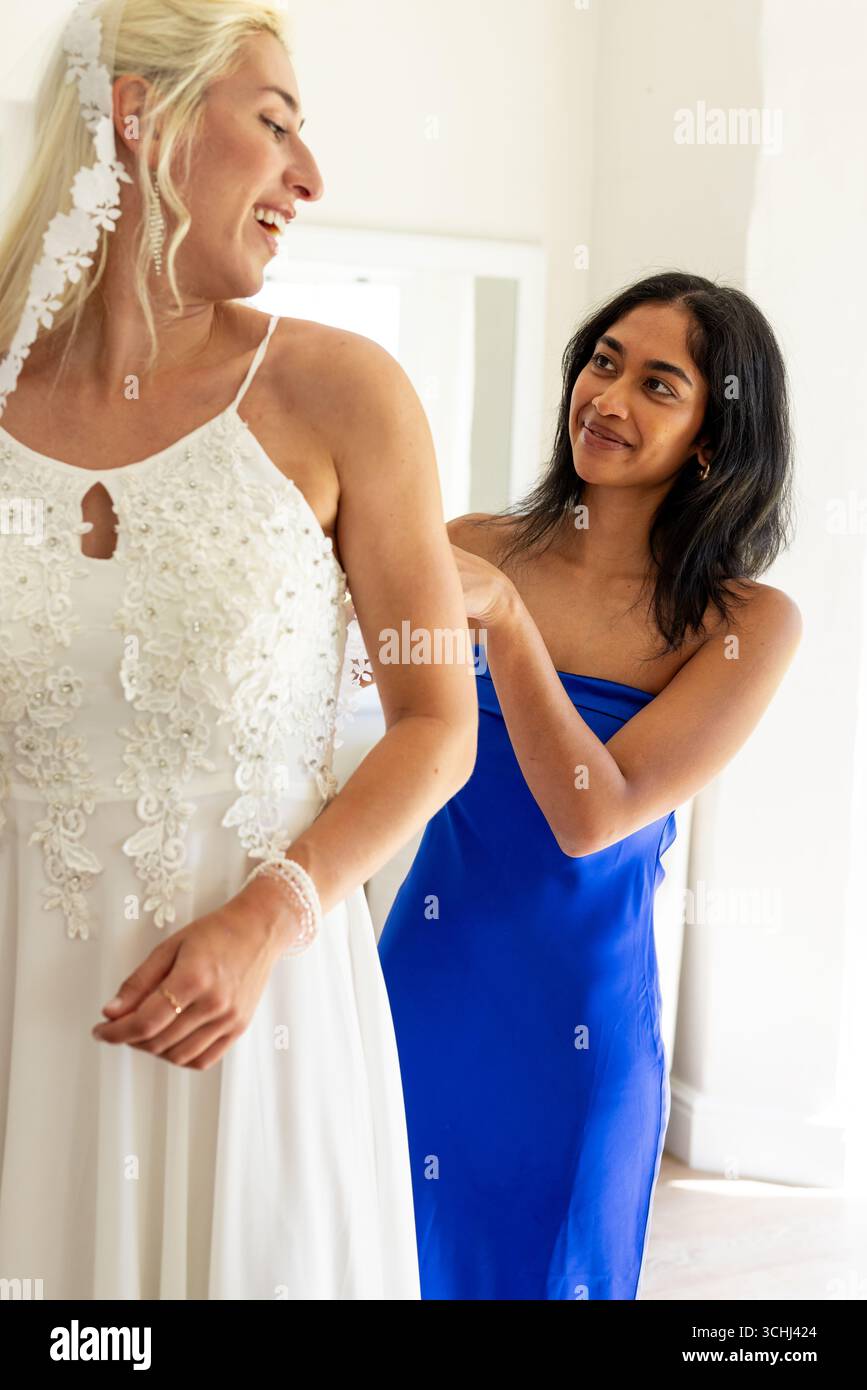 Sposa in abito bianco sorridente mentre damigella d'onore in abito blu regola il suo velo Foto Stock