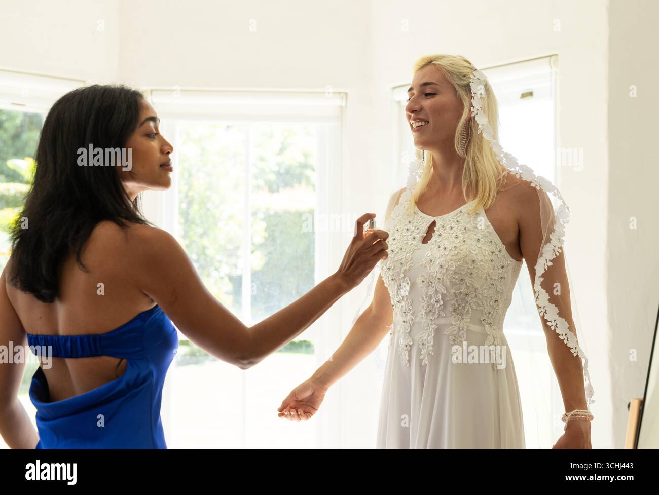 Sposa in abito elegante sorridente mentre la damigella d'onore spruzza profumo in una stanza luminosa Foto Stock