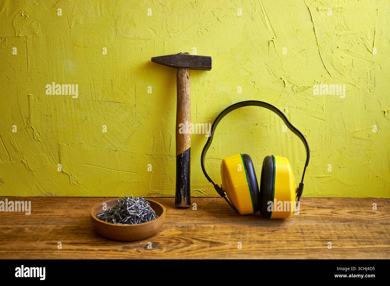 STILL Life con utensili da costruzione: Martello, chiodi in una ciotola di legno e cuffie protettive su uno sfondo giallo a trama. Concetto di costruzione, Foto Stock