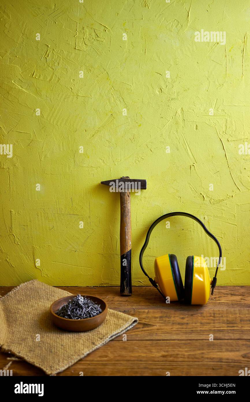 STILL Life con utensili da costruzione: Martello, chiodi in una ciotola di legno e cuffie protettive su uno sfondo giallo a trama. Concetto di costruzione, Foto Stock