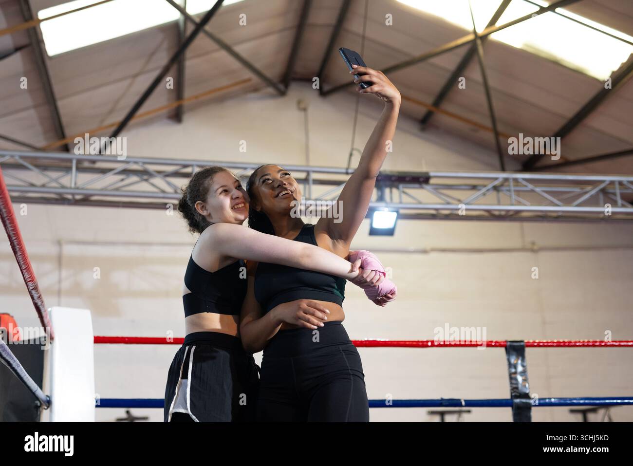 Diverse amiche che scattano selfie in un anello di boxe in palestra con smartphone, corde, impacchi a mano Foto Stock