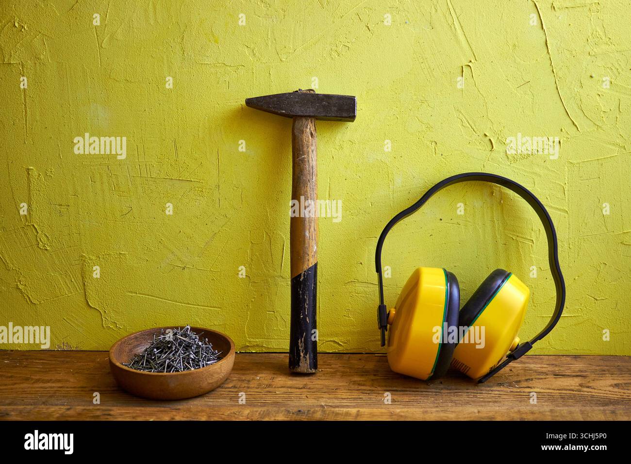 STILL Life con utensili da costruzione: Martello, chiodi in una ciotola di legno e cuffie protettive su uno sfondo giallo a trama. Concetto di costruzione, Foto Stock