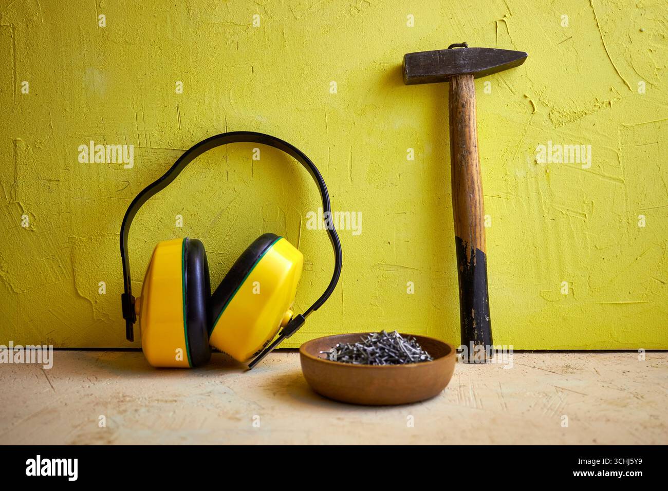 STILL Life con utensili da costruzione: Martello, chiodi in una ciotola di legno e cuffie protettive su uno sfondo giallo a trama. Concetto di costruzione, Foto Stock