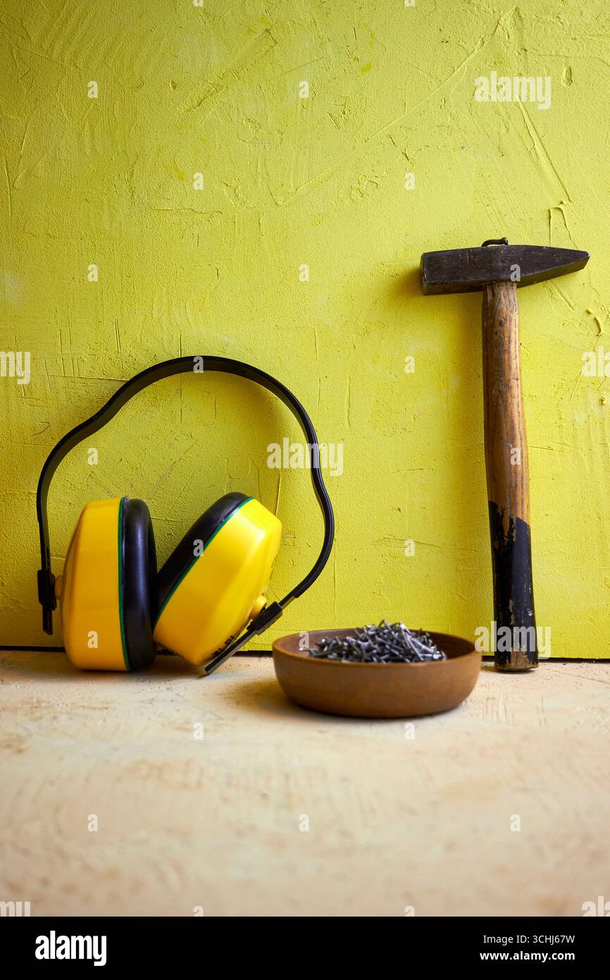 STILL Life con utensili da costruzione: Martello, chiodi in una ciotola di legno e cuffie protettive su uno sfondo giallo a trama. Concetto di costruzione, Foto Stock
