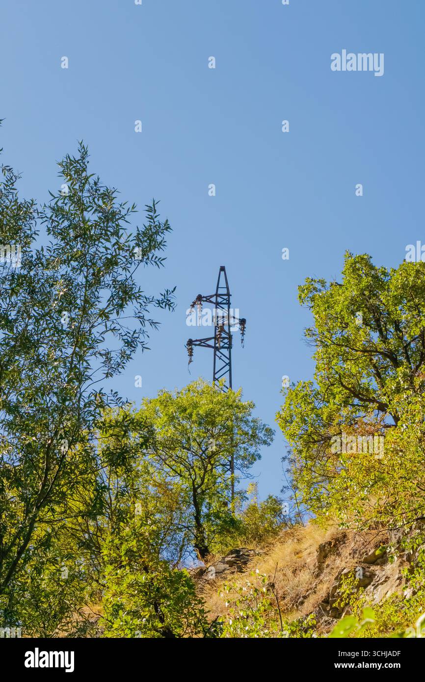 Un'alta torre di trasmissione dell'elettricità sorge su una collina, circondata da alberi verdi vibranti contro un cielo blu brillante, che mette in risalto la potenza essenziale Foto Stock
