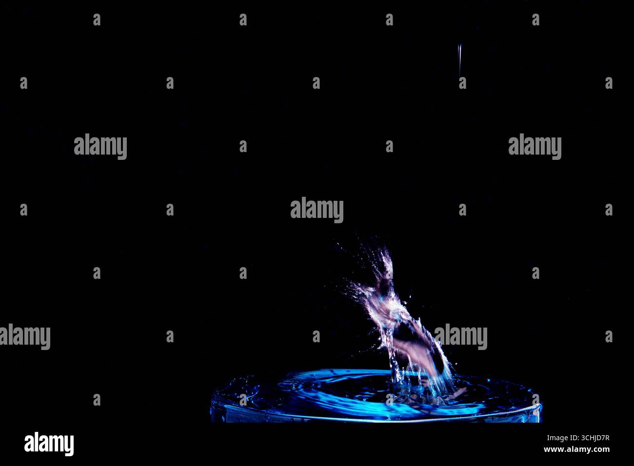 La goccia d'acqua di colore blu colpisce il bordo di vetro, generando uno spruzzo diagonale con increspature su uno sfondo scuro, catturato con tecnica macro ad alta velocità. Foto Stock