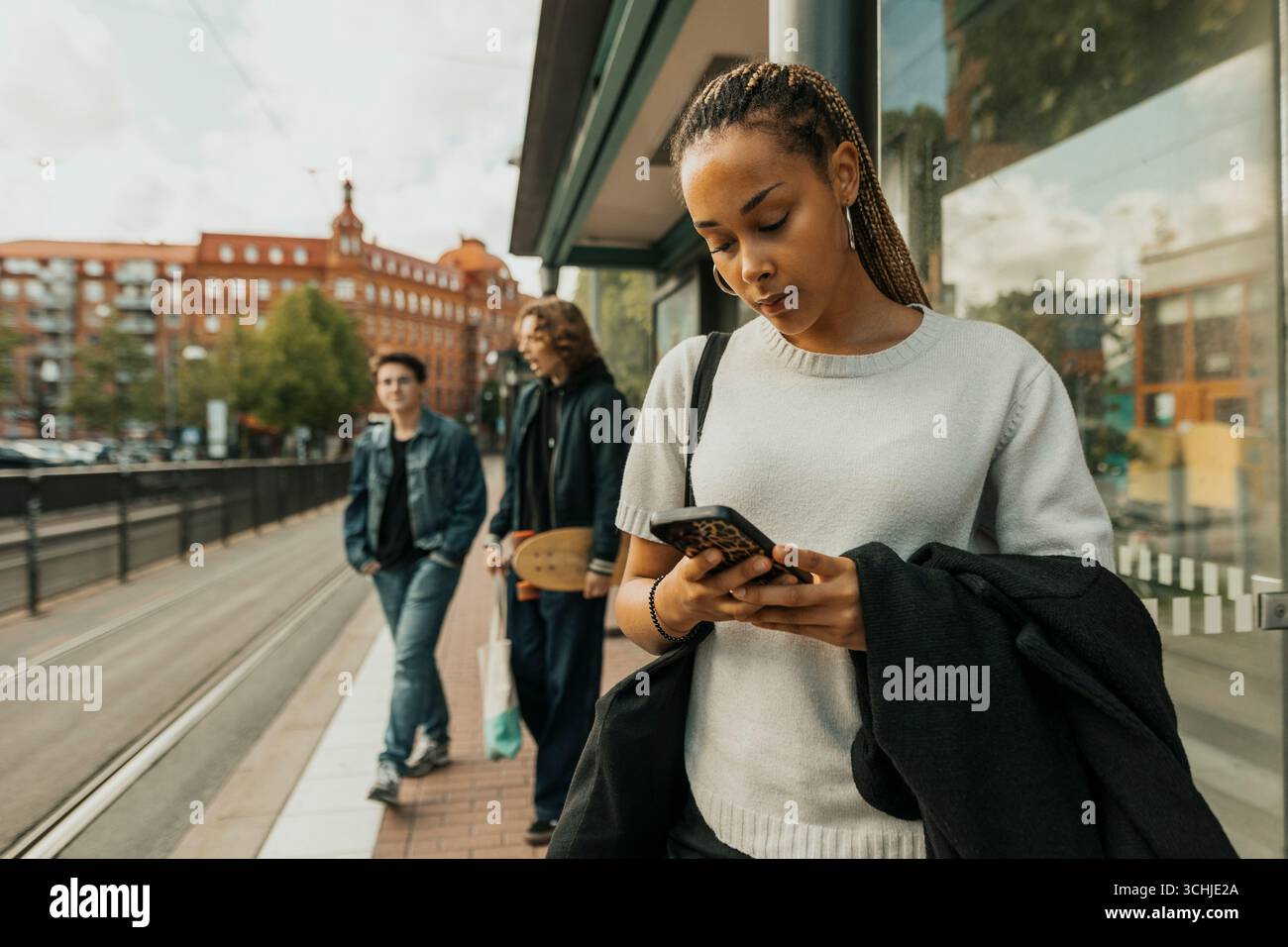 Giovane donna con i capelli intrecciati che usa lo smartphone mentre aspetta alla fermata del tram Foto Stock Giovane donna con i capelli intrecciati che usa lo smartphone mentre aspetta alla fermata del tram Foto Stock