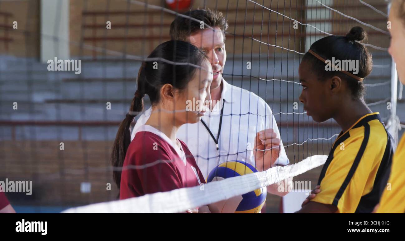 Istruire l'allenatore in gesti di polo bianco ai giocatori che tengono pallavolo nella palestra scolastica, con rete Foto Stock