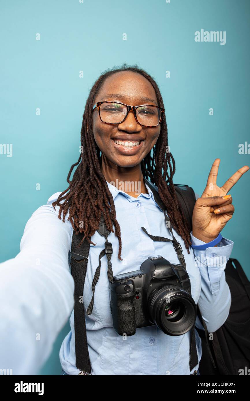 Donna nera eccitata che indossa bretelle e camicia blu, con una reflex digitale intorno al collo, per un gesto V. La fotografa afroamericana si registra in studio, piena di fiducia e positività. Foto Stock