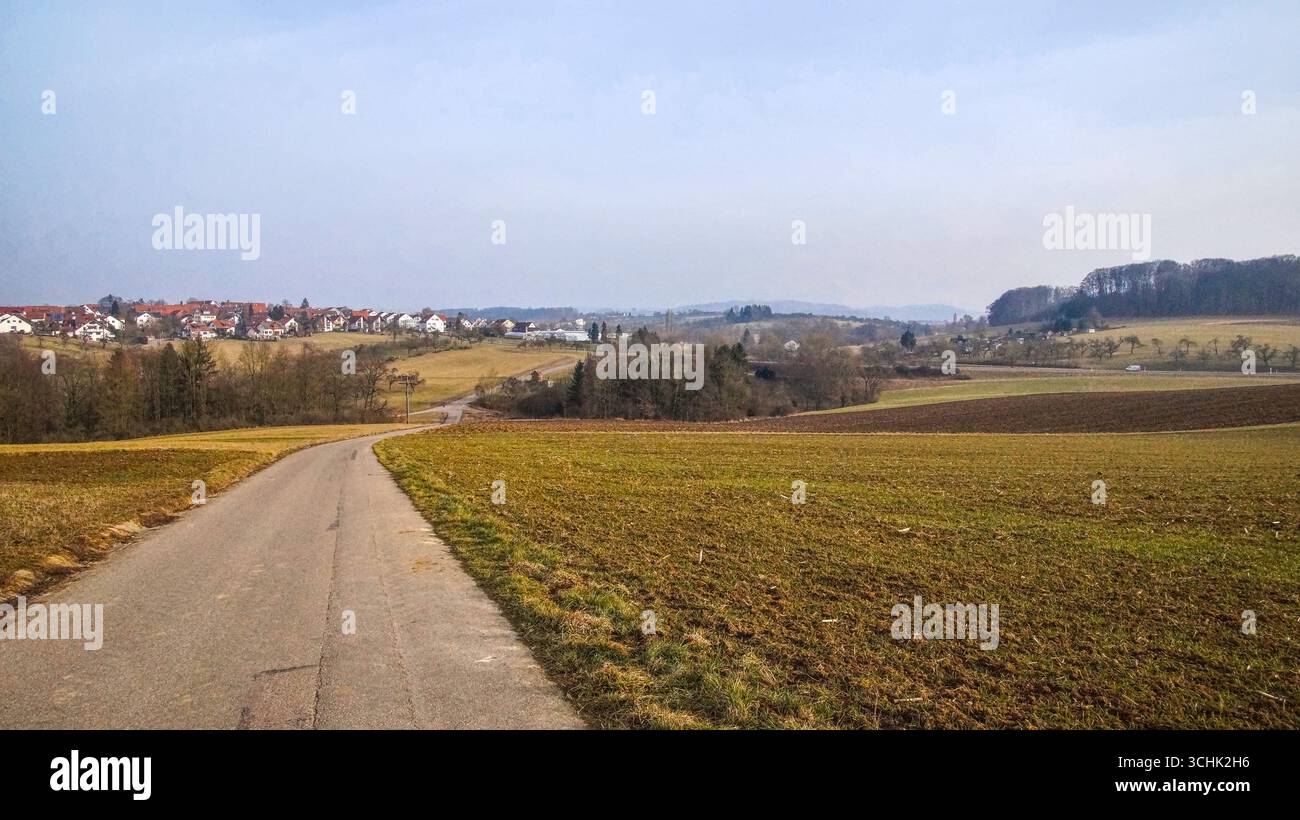 Vista lungo un sentiero rurale sterrato con campi che mostrano la prima crescita verde, che conduce verso un piccolo villaggio sullo sfondo. Foto Stock