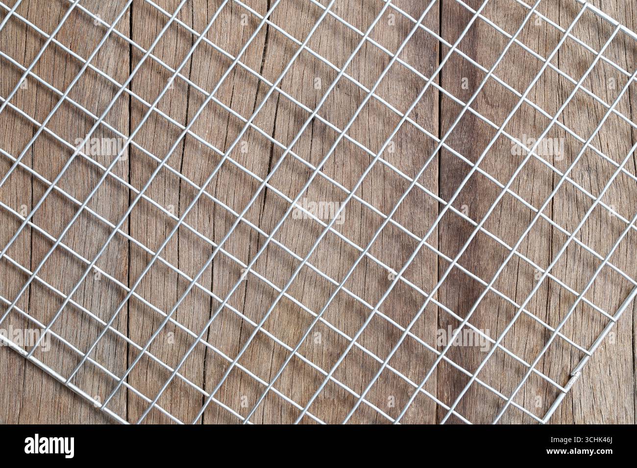 Vista ravvicinata di una griglia a rete metallica quadrata su una superficie in legno, che mostra un mix di texture unico adatto a vari progetti creativi e backgroun Foto Stock