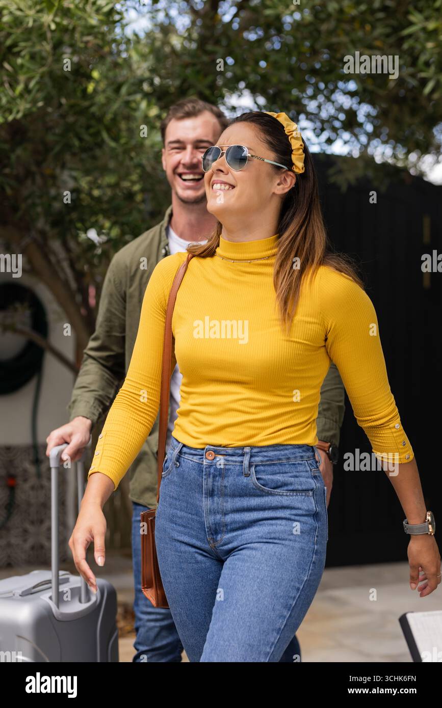 Coppia che si diverte in vacanza, camminando all'aperto con la valigia, sorridendo sotto il cielo soleggiato Foto Stock
