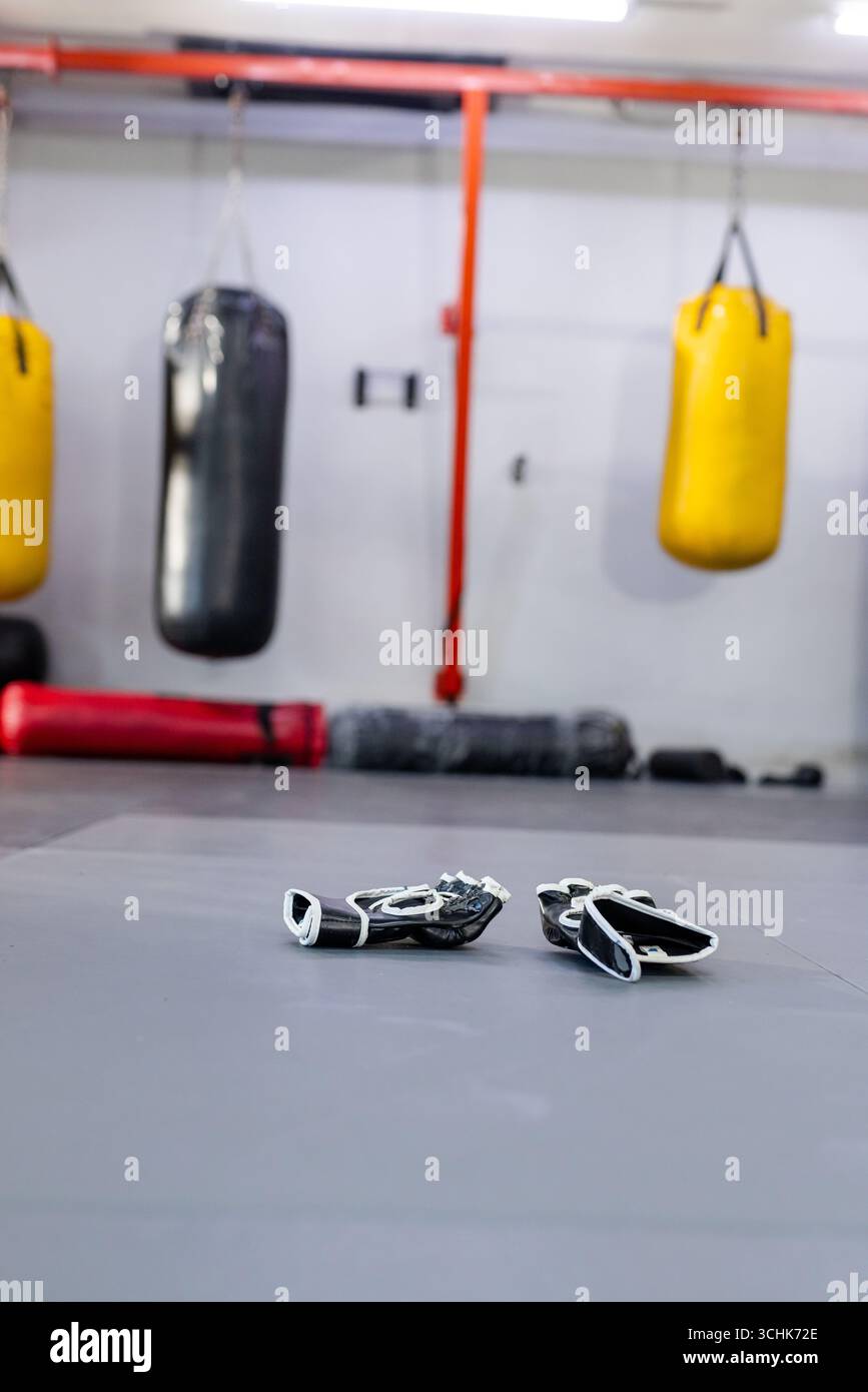 Guanti da combattimento in bianco e nero appoggiati su un tappetino imbottito in palestra, con sacchetti da pugno appesi Foto Stock
