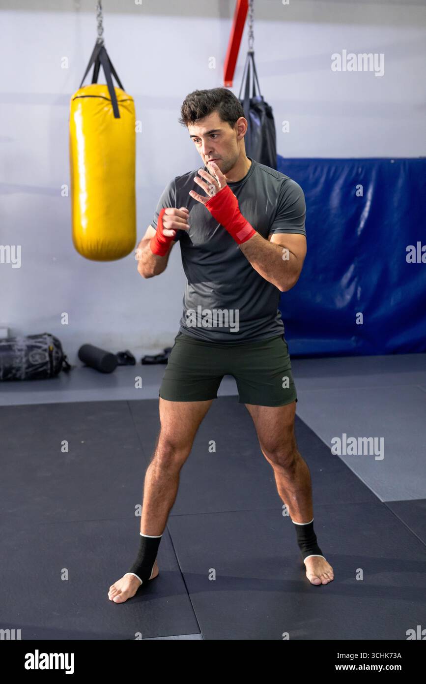 Pugile maschile in piedi in posizione boxe sul tappeto in palestra con sacchetto di punzonatura giallo Foto Stock