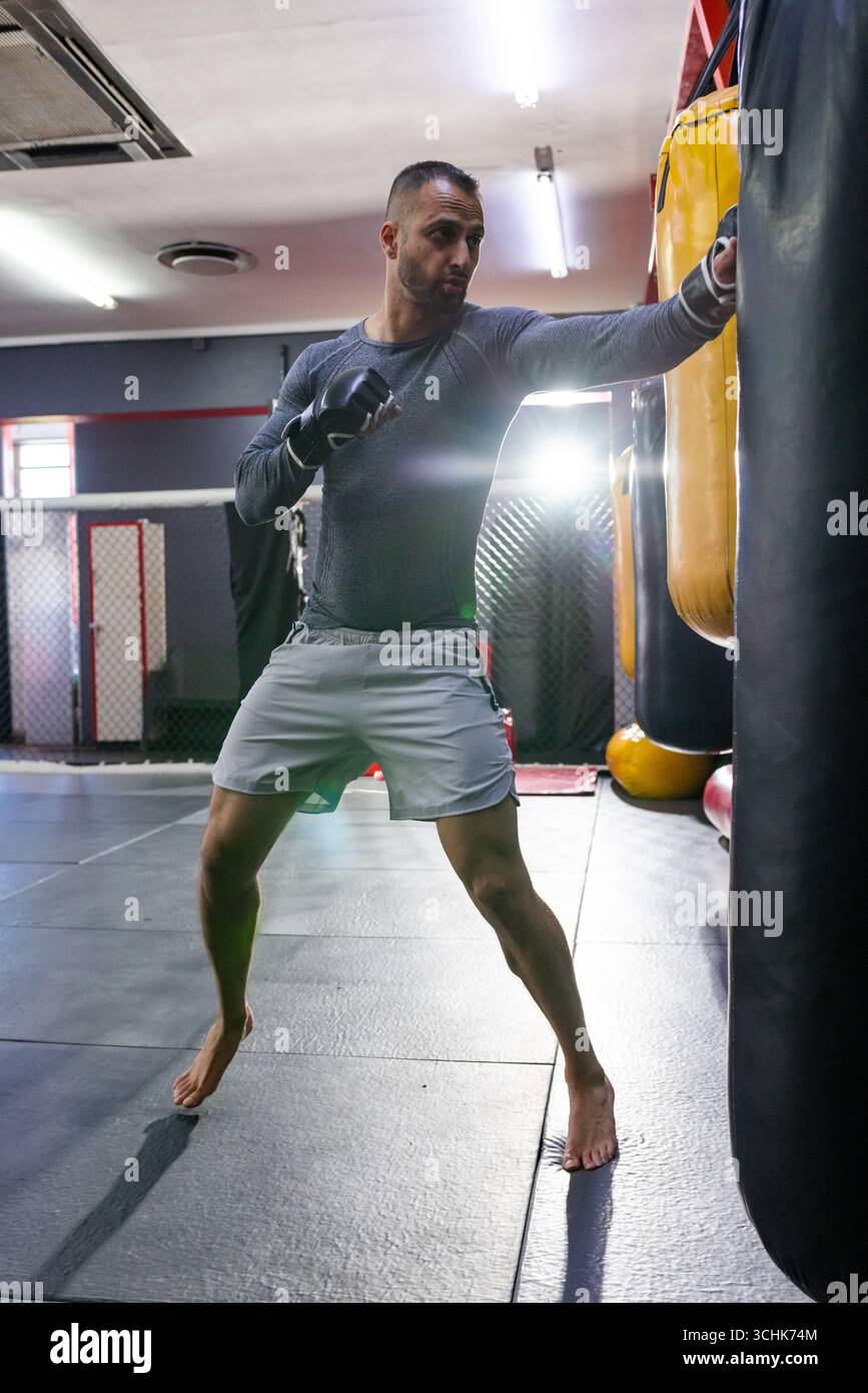 Pugile indiano asiatico medio adulto che indossa la parte superiore su un pavimento imbottito che colpisce la borsa pesante accanto alla gabbia mma Foto Stock