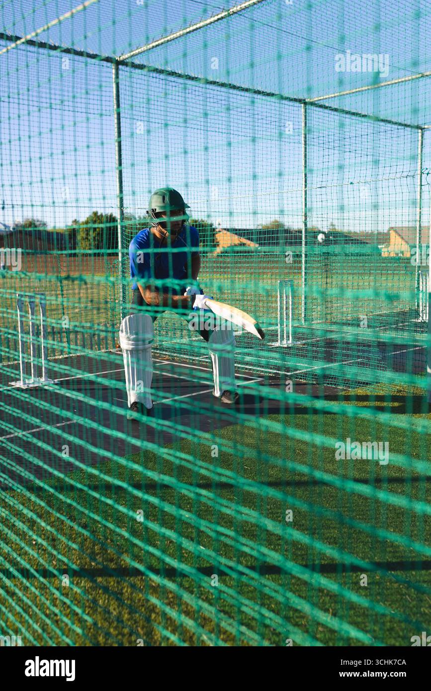 Uomo in piedi in una gabbia di rete di allenamento verde nel casco e pampere che afferrano la mazza di cricket ai ceppi Foto Stock