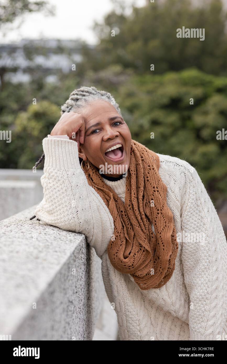 Donna afroamericana anziana appoggiata su ringhiere in pietra nel parco con maglione e sciarpa, ridendo Foto Stock