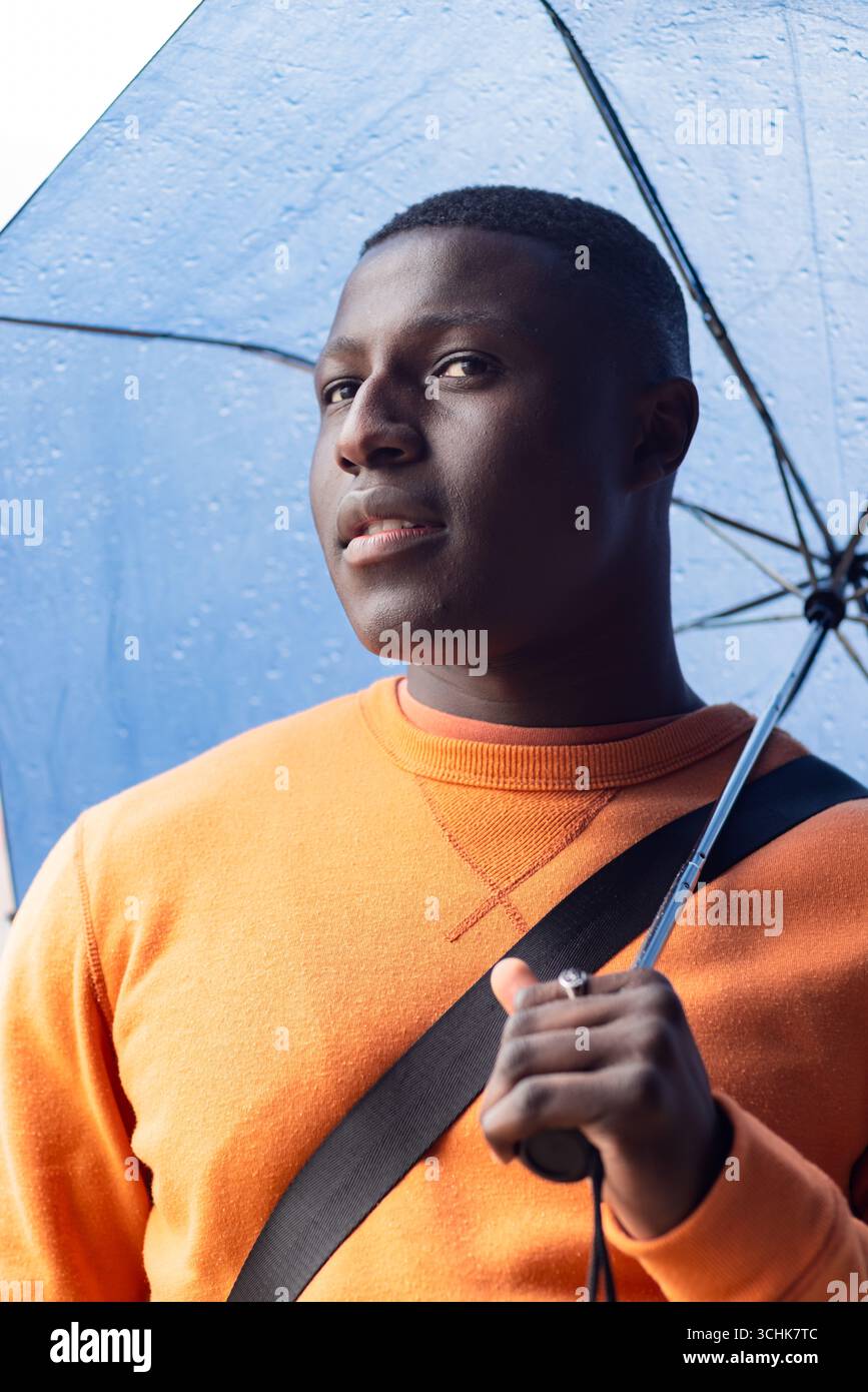 Uomo afro-americano che tiene l'ombrello in testa mostrando il cinturino della borsa e l'anello d'argento sulla strada piovosa Foto Stock