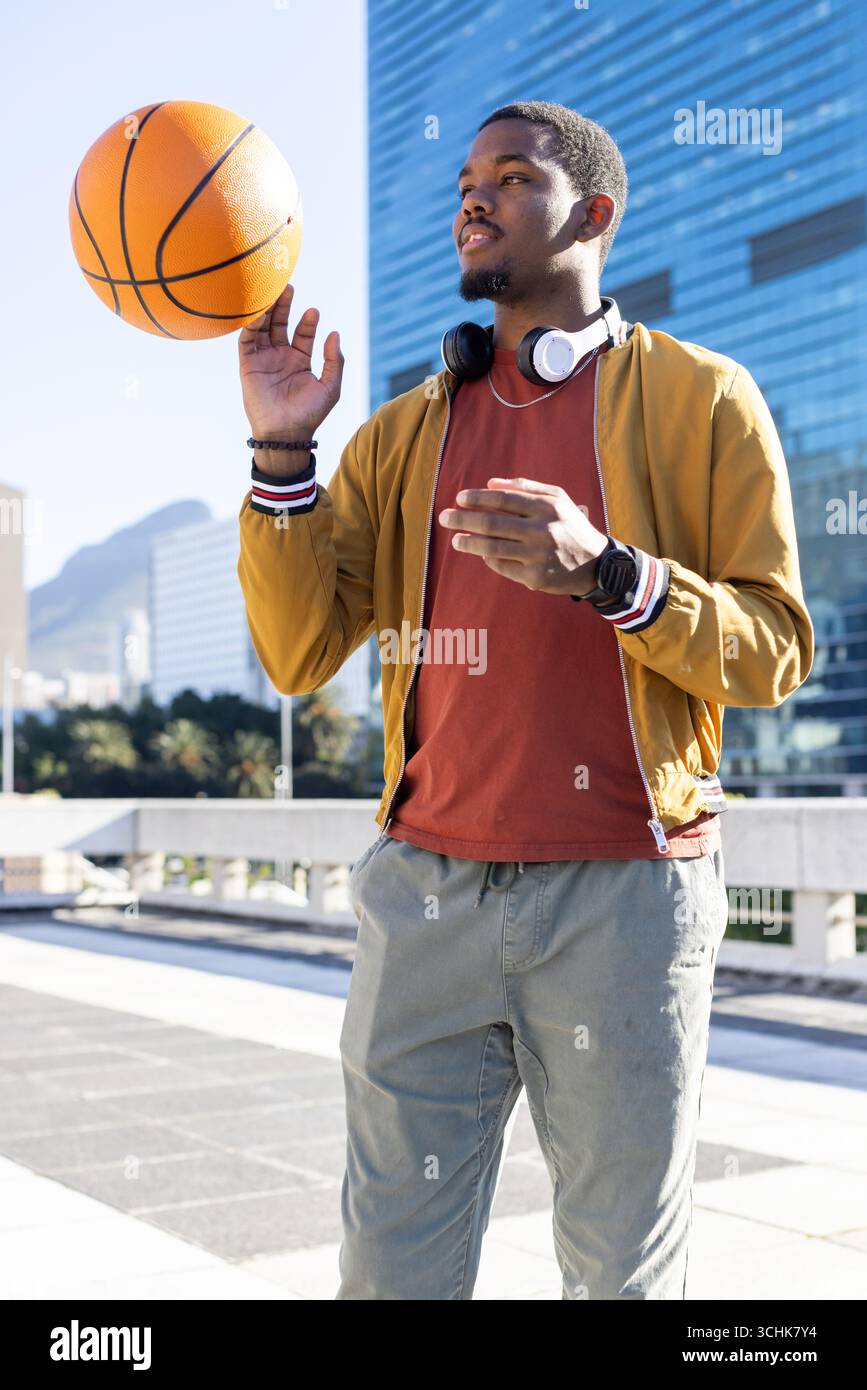 Uomo afroamericano sul tetto che gira da basket su fingertip, bomber, smartwatch Foto Stock