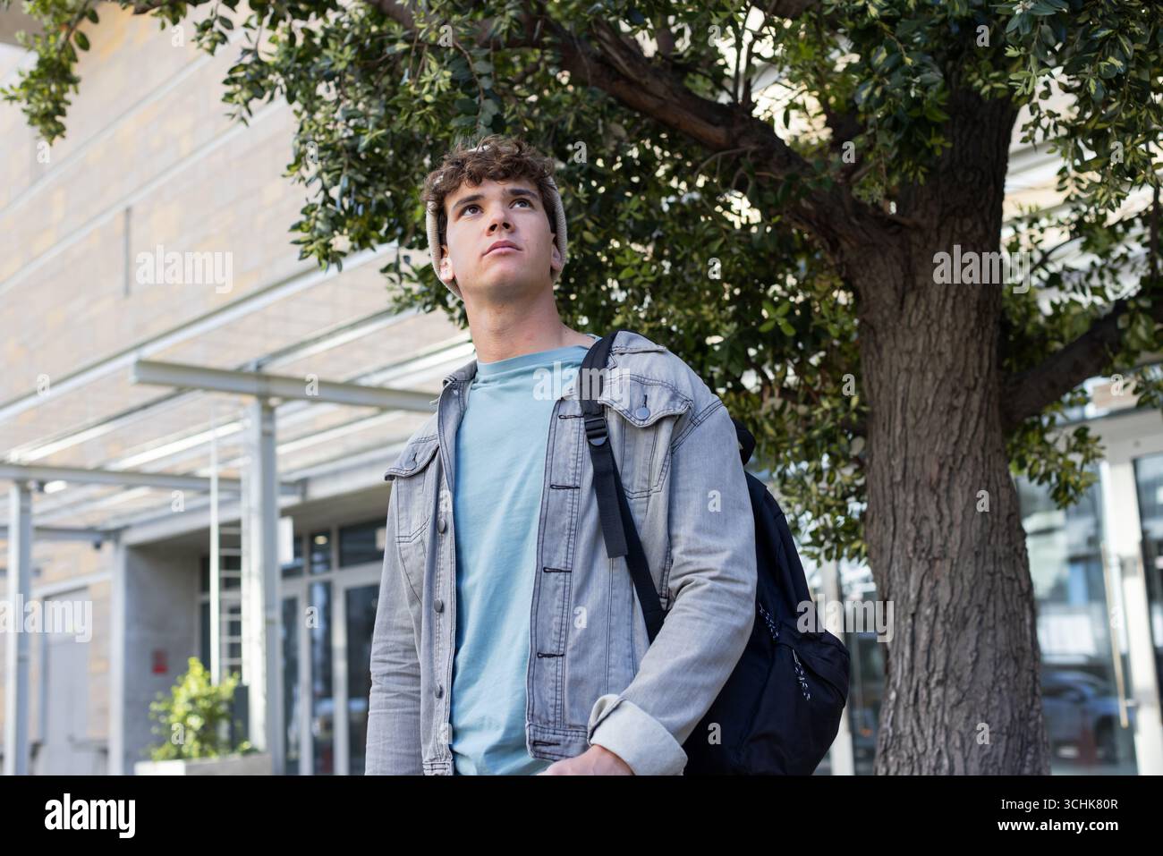 Studente maschio che fa una pausa sotto l'albero davanti a un ingresso moderno che indossa un berretto in maglia, portando con sé uno zaino scuro Foto Stock