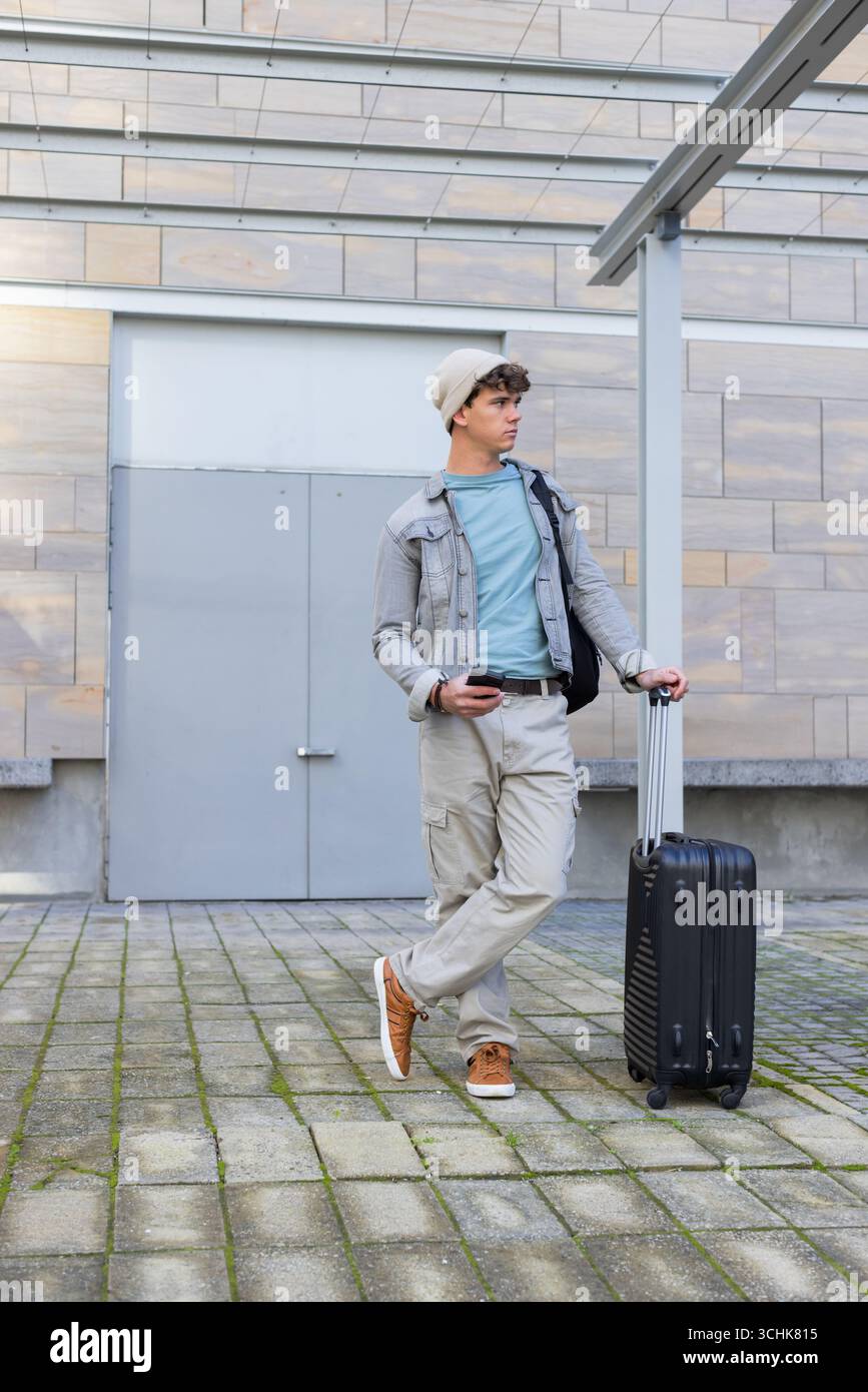 Viaggiatore maschile appoggiato alla maniglia della valigia rotolante su plaza, con smartphone e zaino Foto Stock