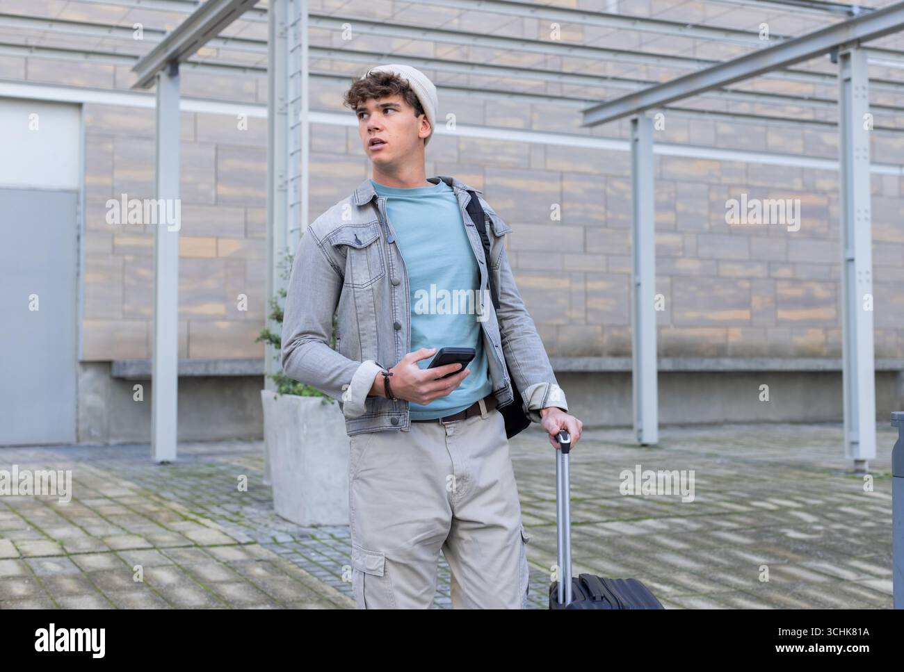 Uomo che tiene in mano smartphone e valigia, che indossa una borsa a spalla sotto la tettoia nel punto di transito Foto Stock Uomo che tiene in mano smartphone e valigia, che indossa una borsa a spalla sotto la tettoia nel punto di transito Foto Stock
