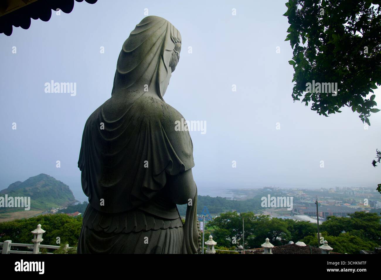 Da una terrazza al Tempio di Bomunsa, un Bodhisattva in pietra si affaccia sulla costa di Sagye e sui quartieri costieri di Seogwipo, con alberi, tetti e Foto Stock