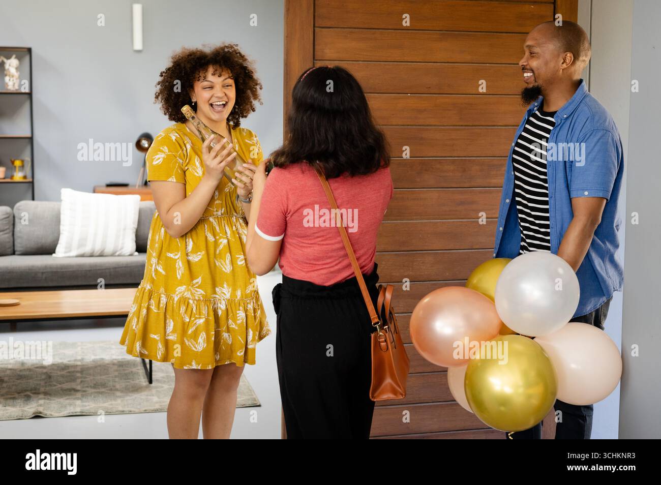 Alla festa, amici diversi che celebrano una festa a sorpresa a casa, tengono palloncini e ridono gioiosamente Foto Stock