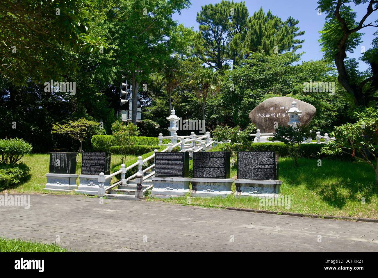 Un prato terrazzato al parco Cheonjeyeon mostra targhe nere interpretative e un grande masso inciso accanto a lanterne di pietra e ringhiere tra manicure Foto Stock