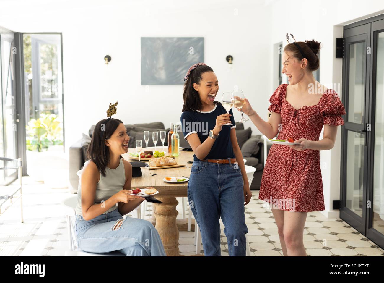 Ridendo e brindando con bicchieri da vino, tre giovani donne si godono il pomeriggio soleggiato all'interno Foto Stock