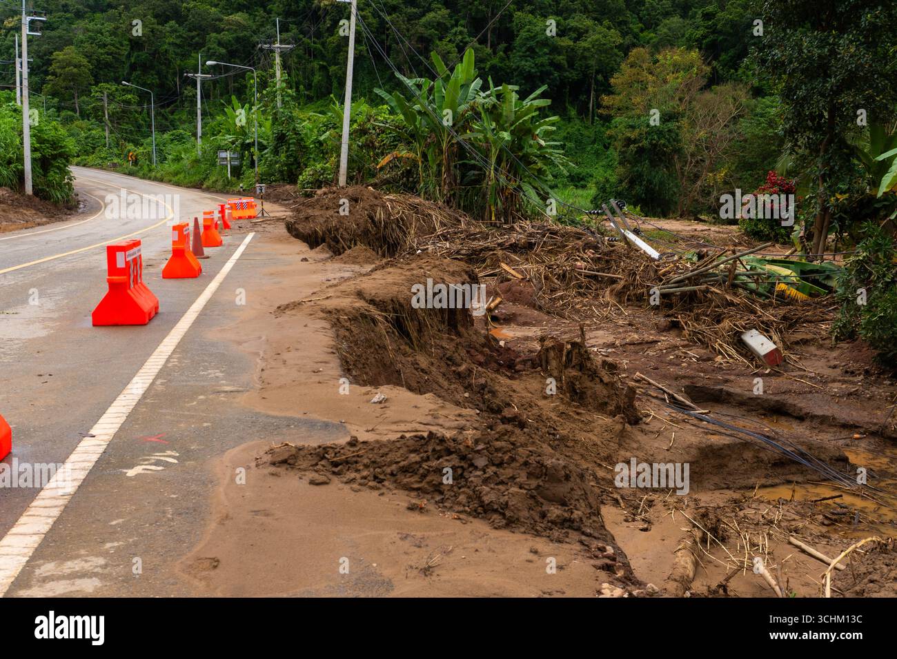 Strada rurale gravemente danneggiata da inondazioni e frane, con fango, erosione del suolo e barriere arancioni posizionate per sicurezza e avvertenza. Foto Stock