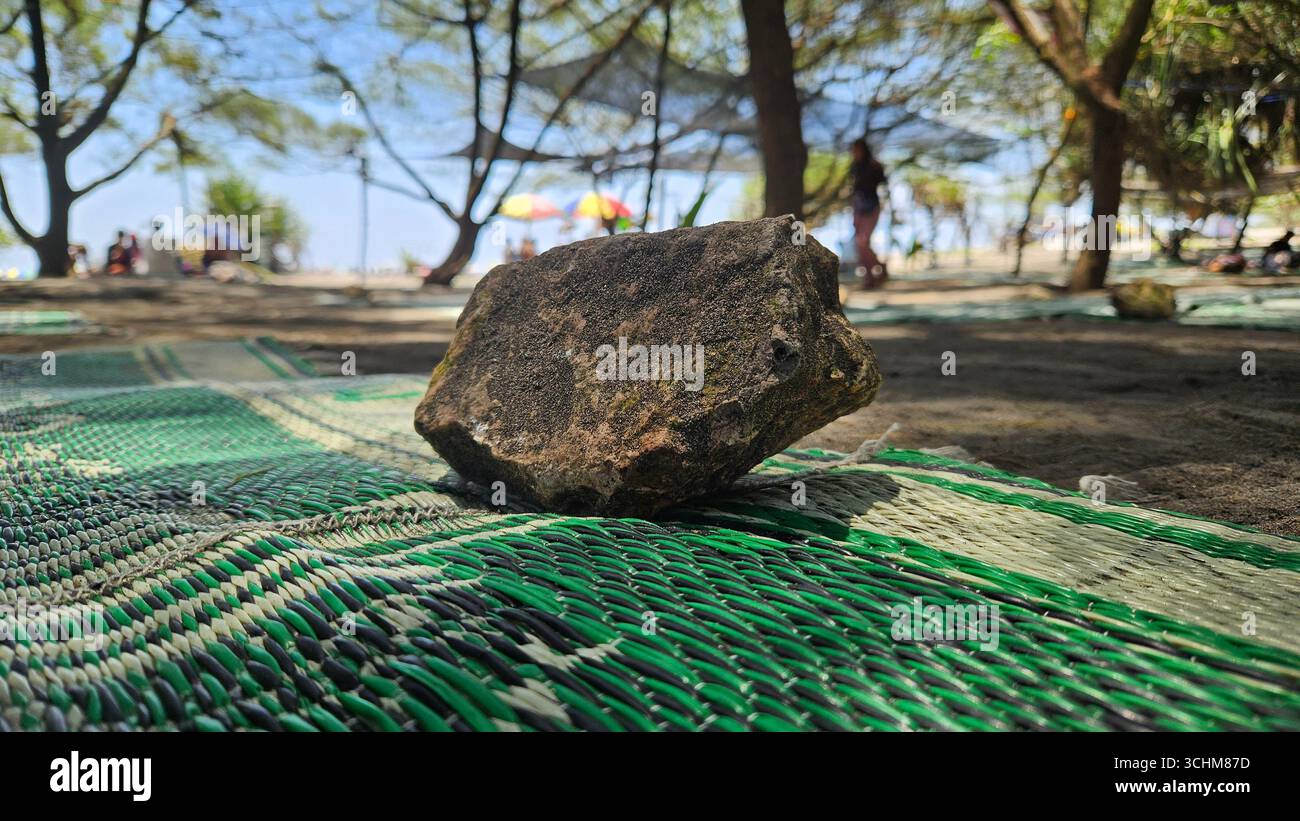 Roccia collocata su un tappeto di tessuto verde in un ambiente esterno con alberi e persone sullo sfondo, dettaglio in pietra naturale - Immagine stock catturata con smartphone