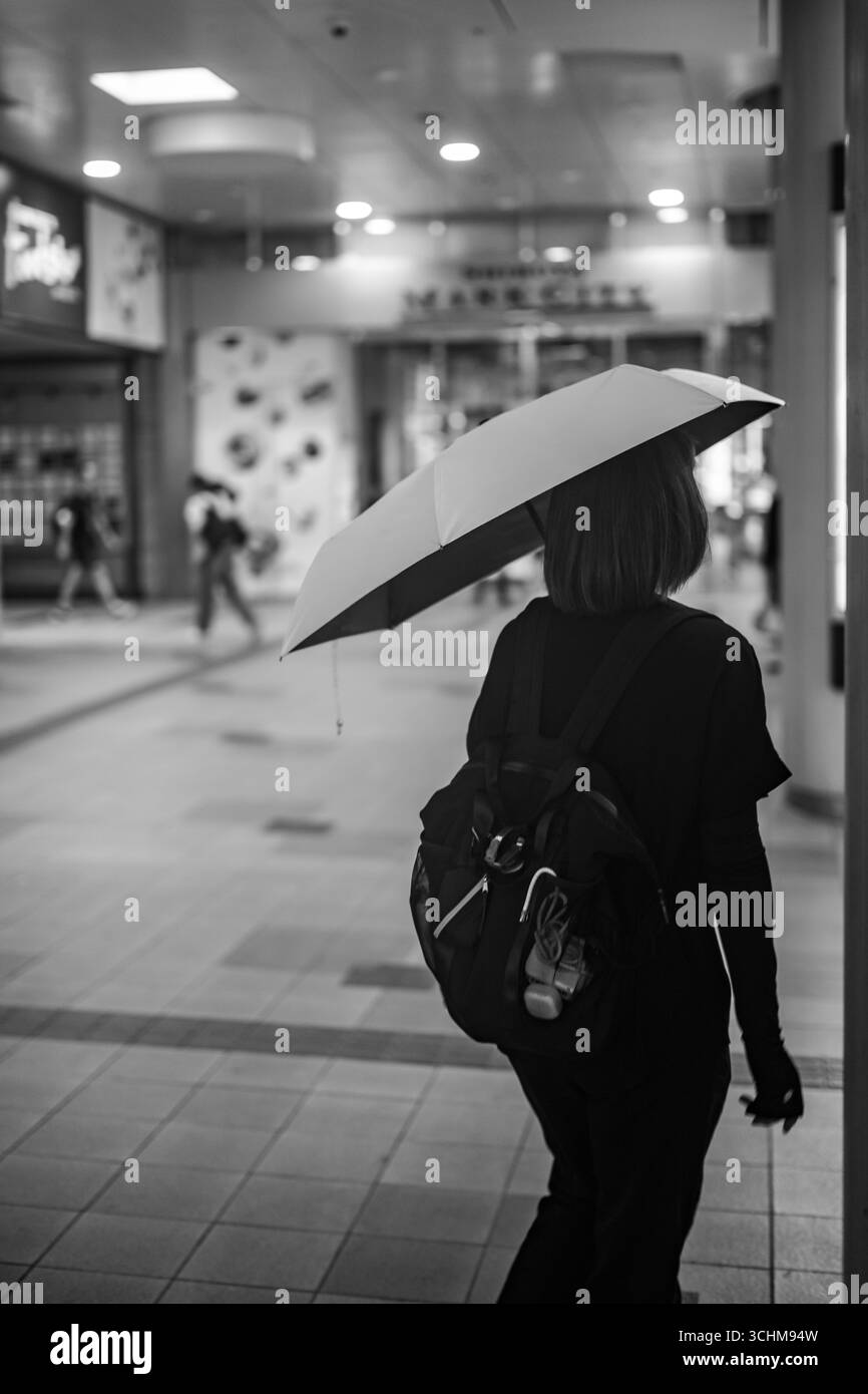 La donna sta da sola sotto la pioggia tenendo in mano un ombrello, colta in un momento di calma forza e riflesso. I toni monocromatici aumentano l'emozione Foto Stock