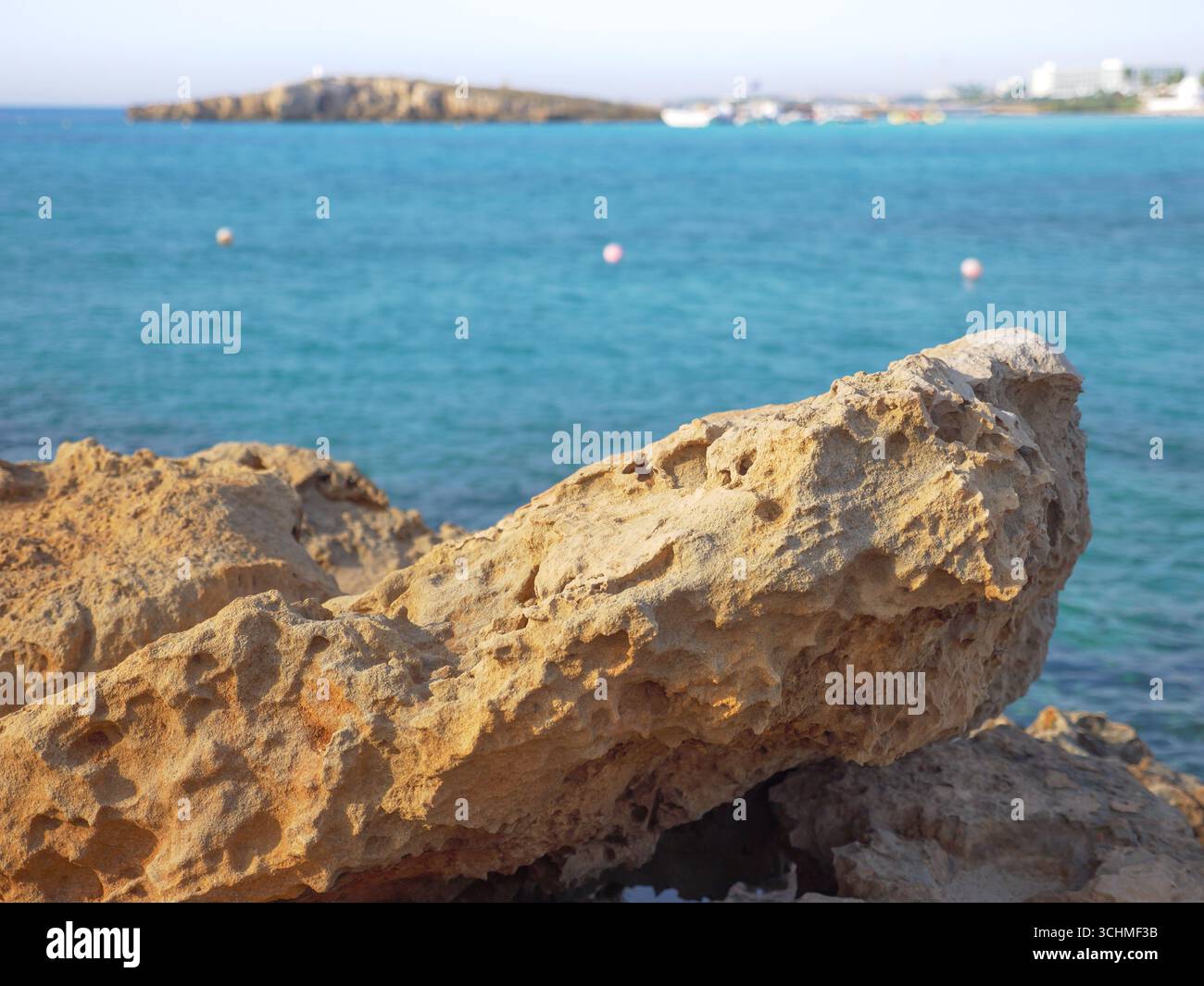 Vista da dietro di una grande roccia porosa gialla su uno splendido mare Mediterraneo blu, la spiaggia di Nissi, una piccola isola e gli edifici alberghieri sullo sfondo. Foto Stock