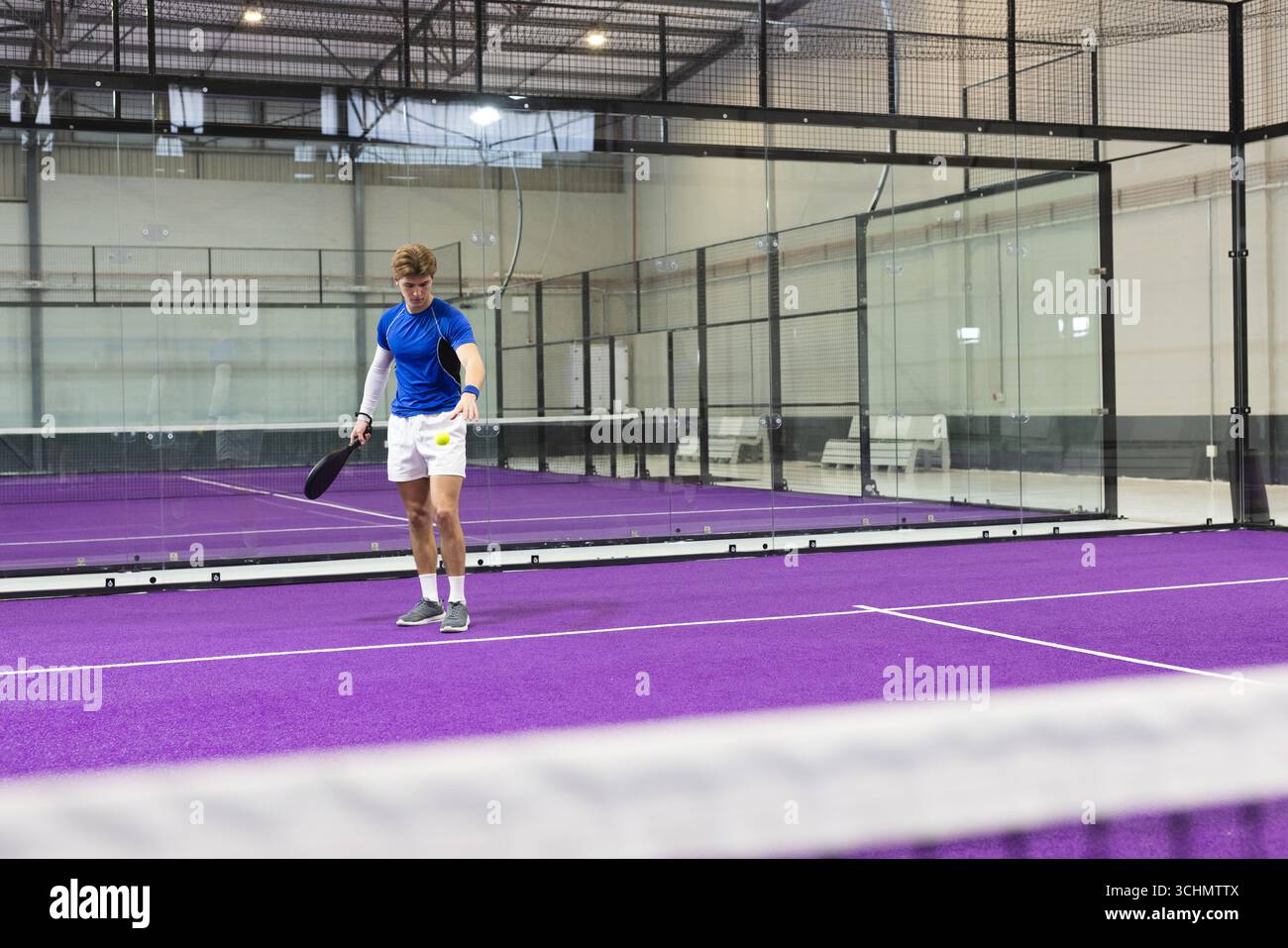 Giocatore di padel maschile in piedi all'interno del campo di padel di vetro con racchetta accanto alla palla da tennis gialla Foto Stock