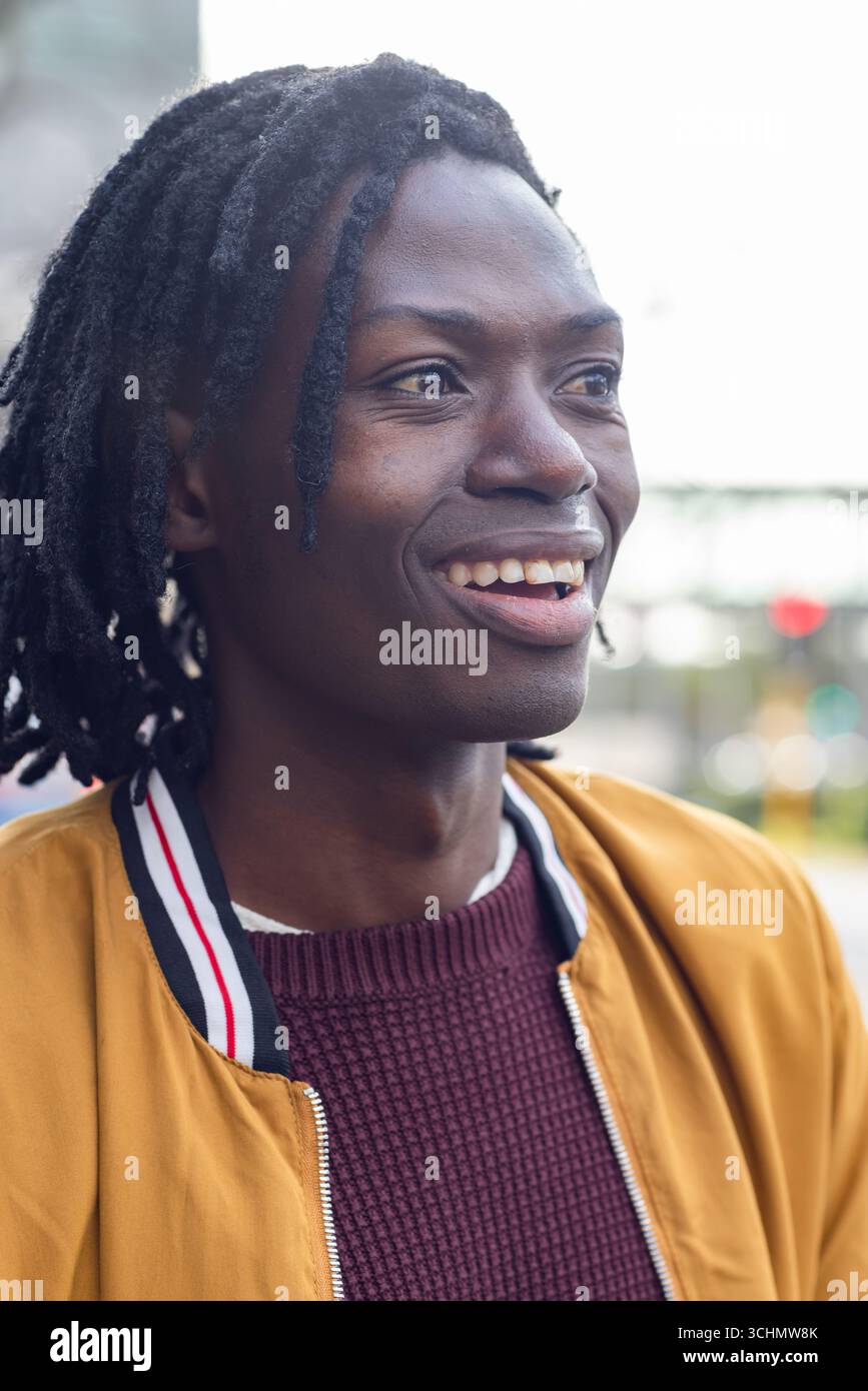 Uomo afro-americano sorridente mentre si trova in città e indossa un bomber e un maglione in maglia Foto Stock