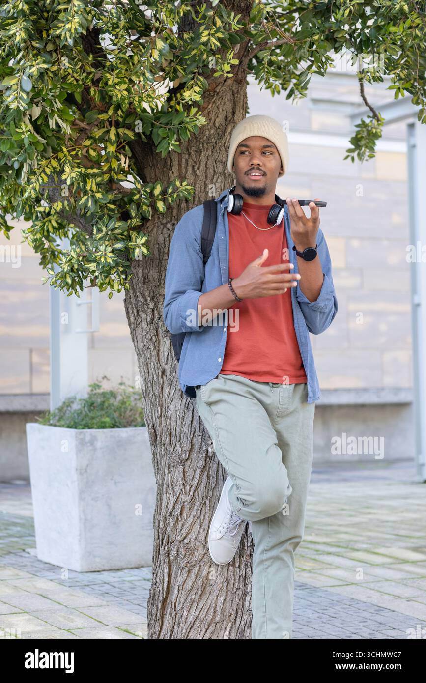 Uomo afro-americano appoggiato contro un albero in piazza con smartphone e cuffie in mano Foto Stock