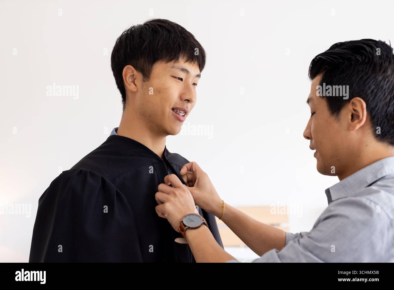 Padre orgoglioso che aiuta il figlio a regolare l'abito da laurea a casa, entrambi sorridenti calorosamente Foto Stock