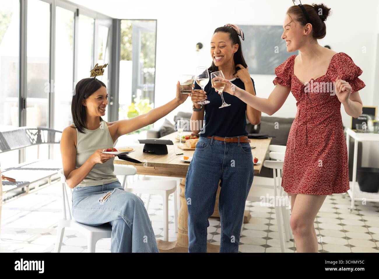 Ridendo e brindando con bicchieri da vino, tre giovani donne si godono il pomeriggio soleggiato all'interno Foto Stock