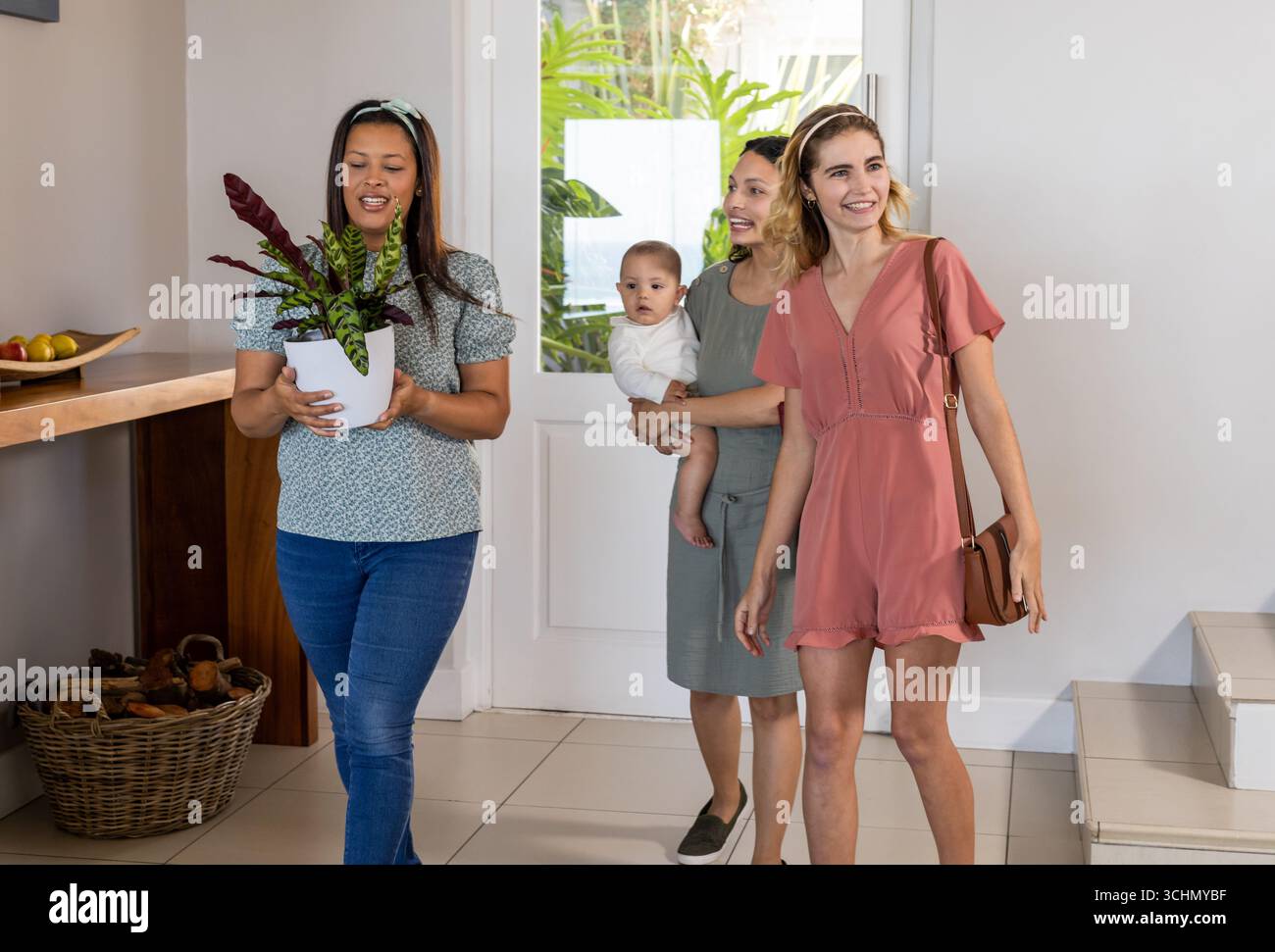Diverse amiche che arrivano a casa, sorridono e trasportano piante, bebè in braccio Foto Stock