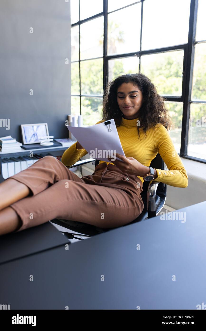 Donna sicura di sé in maglione giallo che esamina documenti con i piedi sulla scrivania, in ufficio e in ufficio Foto Stock