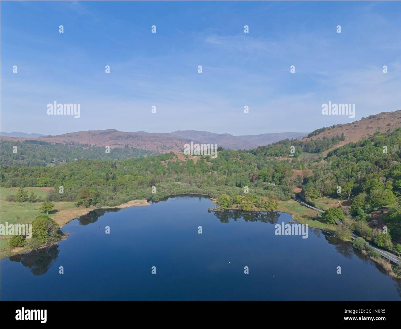 Una vista aerea mostra un tranquillo lago circondato da lussureggianti alberi verdi e colline ondulate sotto un cielo blu luminoso. Foto Stock