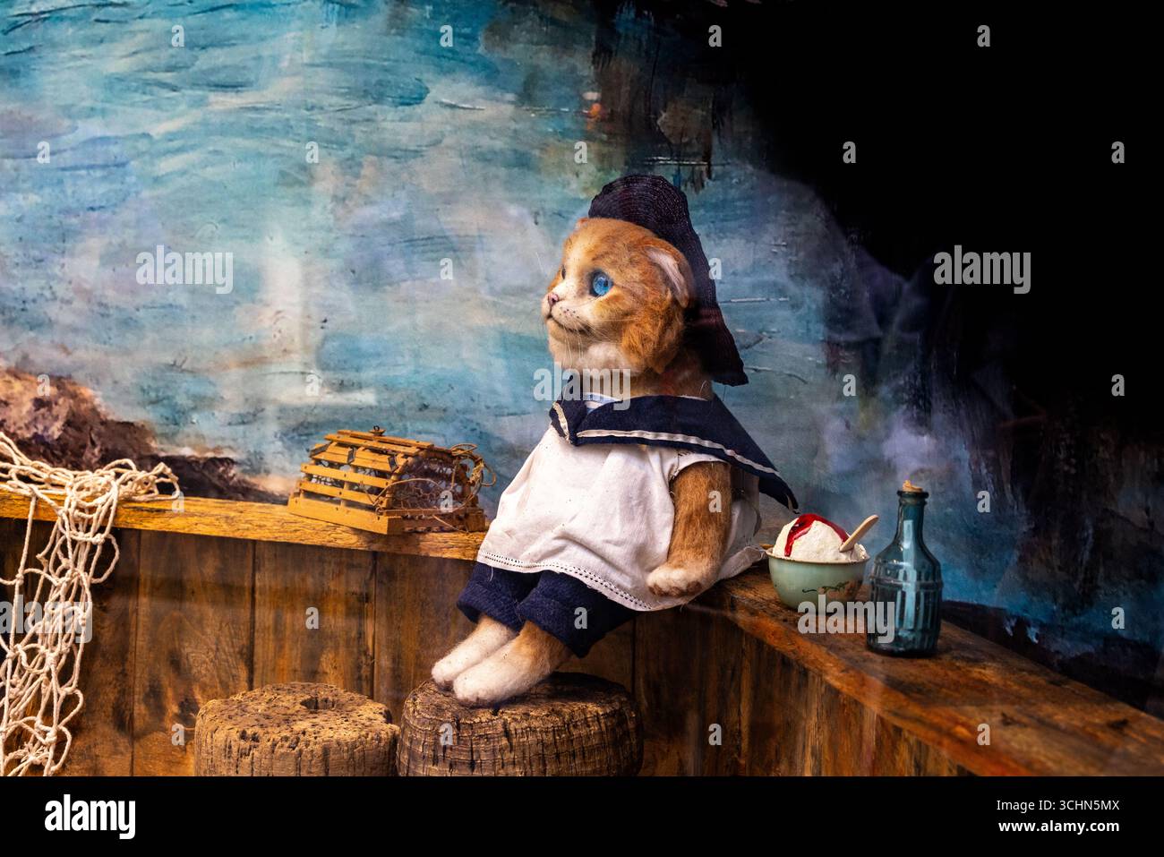 Gatto di peluche in un costume da marinaio seduto su una botte in un'esposizione a tema nautico con rete da pesca, trappola di aragoste e oggetti di scena vintage. Foto Stock