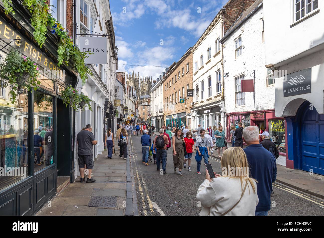 Turisti e gente del posto in una strada trafficata di York, in Inghilterra, con edifici storici, negozi e guglie della cattedrale di York che si innalzano sullo sfondo. Foto Stock